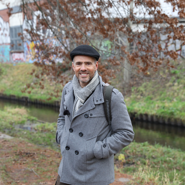 Ein Mann steht warm eingepackt mit Mütze und grauem Mantel auf einer Grünfläche neben dem Kanal.